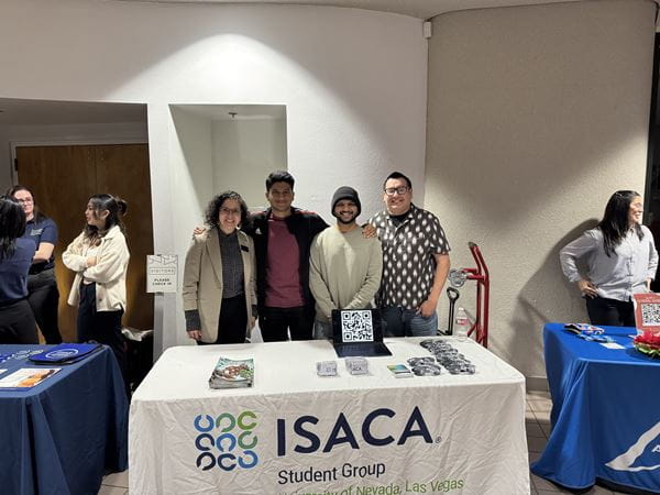 A group of people gathered around a table displaying an ISACA sign, engaged in discussion or collaboration.