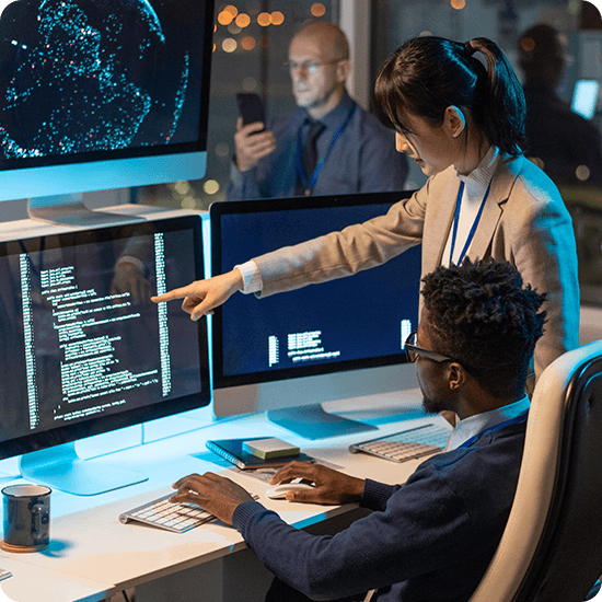 Two people engaged in work on their computers, with a large monitor displaying information in front of them.