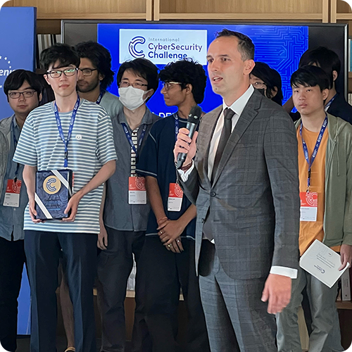 A host at the International Cybersecurity Challenge with a bunch of contestants behind him