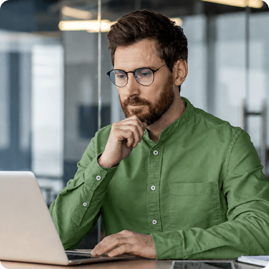A bearded man sitting at a desk, focused on his laptop while working in a well-lit office environment.