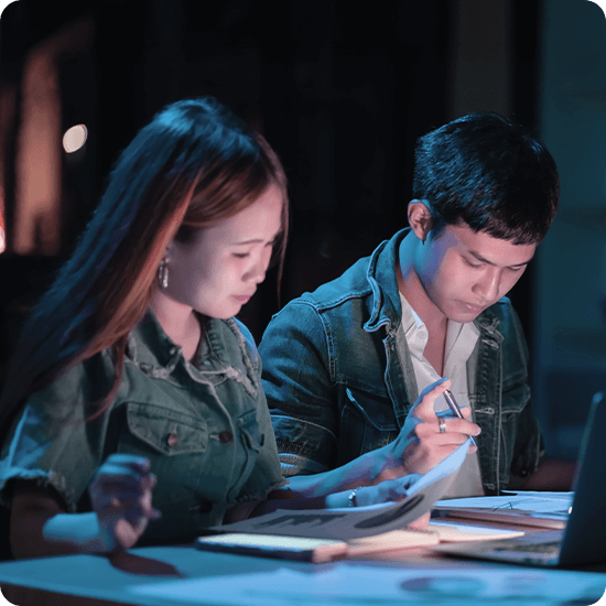 A man and woman sit at a table, focused on a laptop, engaged in discussion or collaboration.