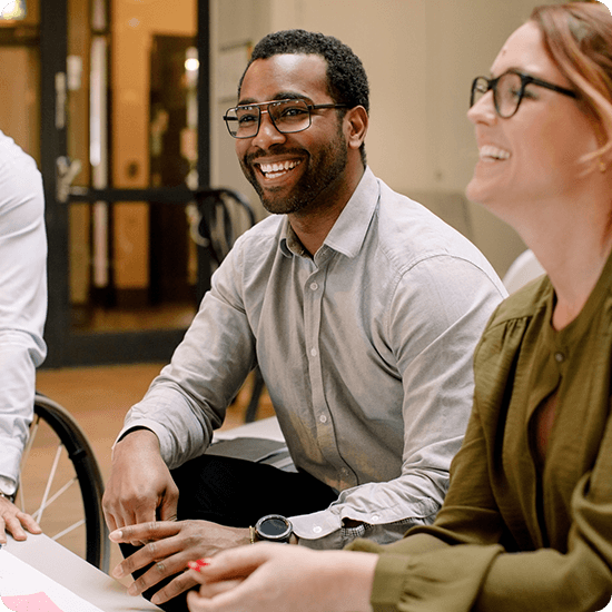 Smiling diverse colleagues, including a Black man and a white woman with glasses, sit together in a bright office, conveying warmth and teamwork.