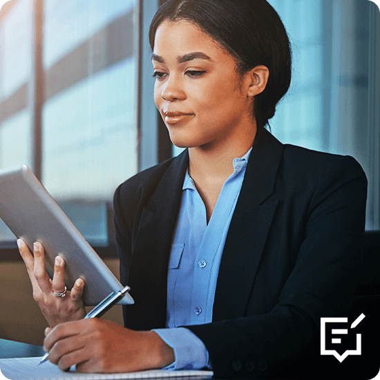 A businesswoman in a suit writes on paper with a tablet in hand, focused on her work at a desk.